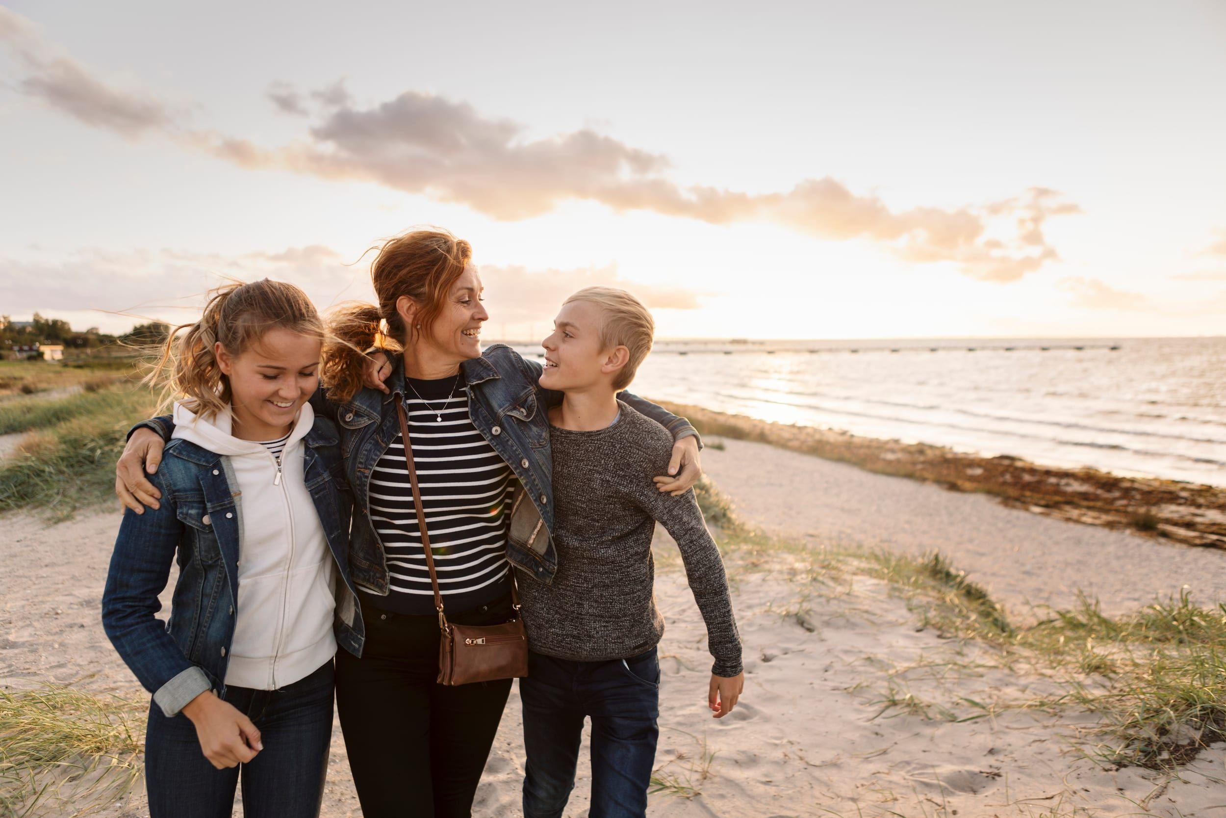 A mother and two teenagers walking arm in arm on a beach in evening light